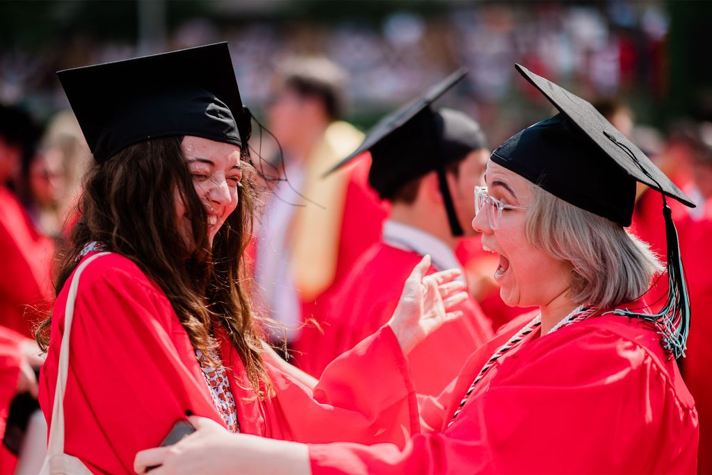 Photo of two young white women looking at each other excitedly. The women on the right has ash blonde hair and holds the arms of the women on the left, who has long brown hair and laughs. Other grads can be seen around them in the blurry background. Everyone wears a red gown and black caps.