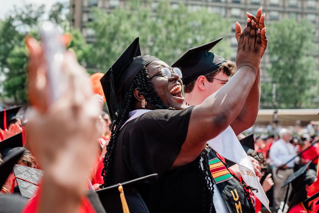 Photo of a young black woman with long, Senegalese twists cheers excitedly and class as she faces an unseen stage. She wears a black dress, black cap, and black sash with Kente stripes on it. Other graduates around her wear red gowns and black caps.