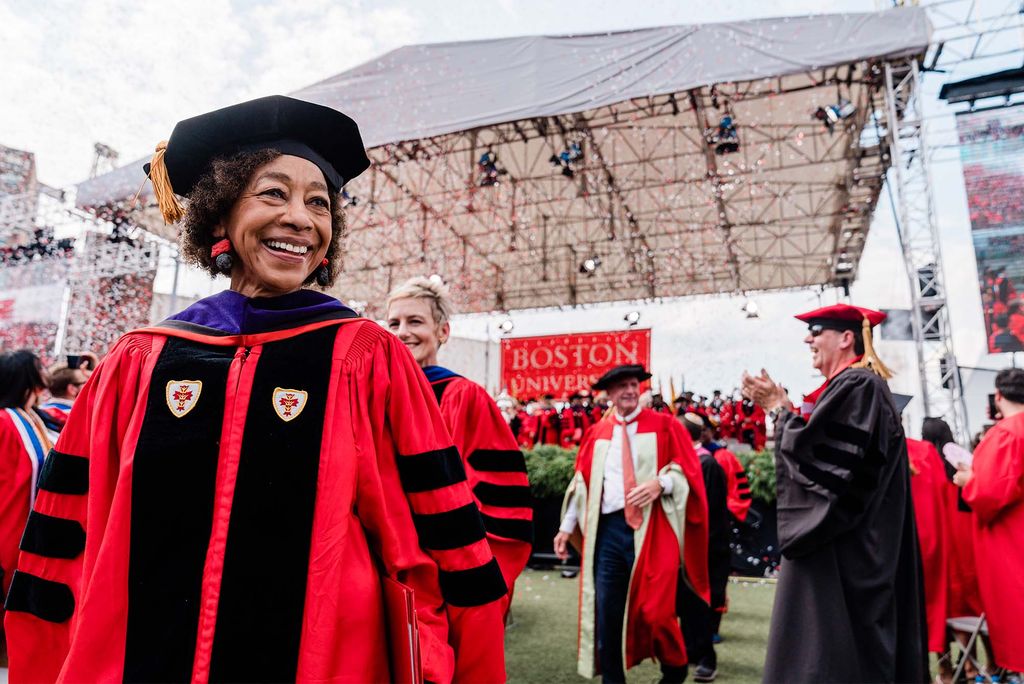 Photo of Gay J. McDougall, at left, smiling, during commencement. She is a middle-aged Black woman who wears a red graduation robe with black stripes, black sash, and black hat. Others are seen processing off of nickerson with her.
