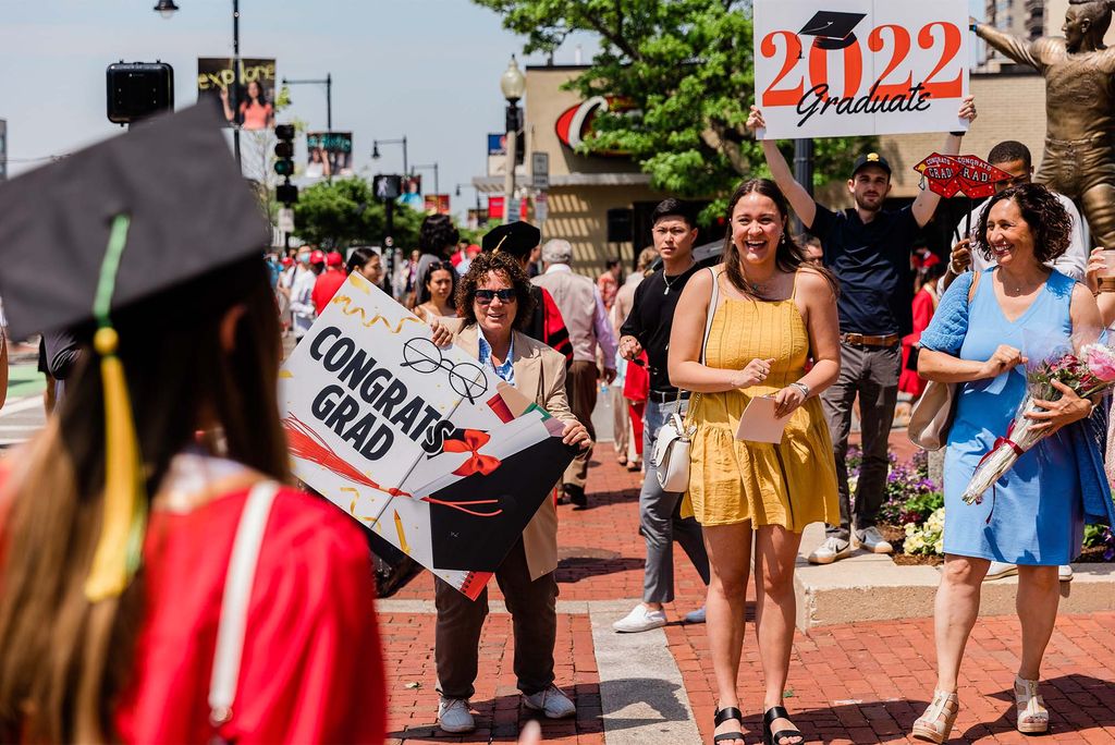 A group of people, possibly a family, stands along the Comm Ave sidewalk holdings that say "Congrats Grad 2022!". At front, are three White women, the middle one wears a yell dress, one at right wears a blue dress. Students processing towards nickerson can be seen in the foreground and background.
