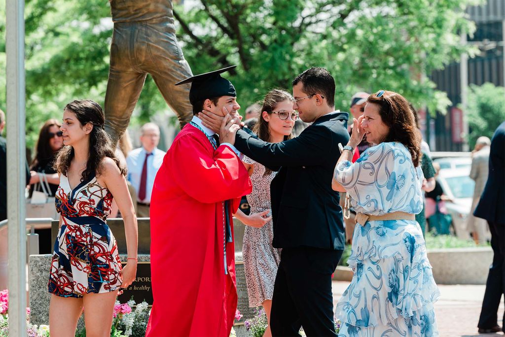 Photo of a young graduate wearing a red gown and black cap having his face embraced by a relative in a black suit. A middle-aged woman beside them wears a light blue dress as a young woman to his left wears a white, red, and blue romper. The legs of a bronze statues can be seen behind them.
