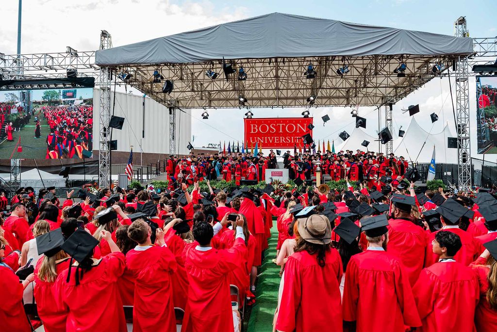 Wide shot photo of the 2022 Boston University Commencement. The stage with speakers, honorees, ands deans wearing black and red robes can be seen front and center as a jumbotron i seen to the left showing the scene on stage. Students wearing red graduation gowns begin to throw black caps into the air from their seats in front of the stage.