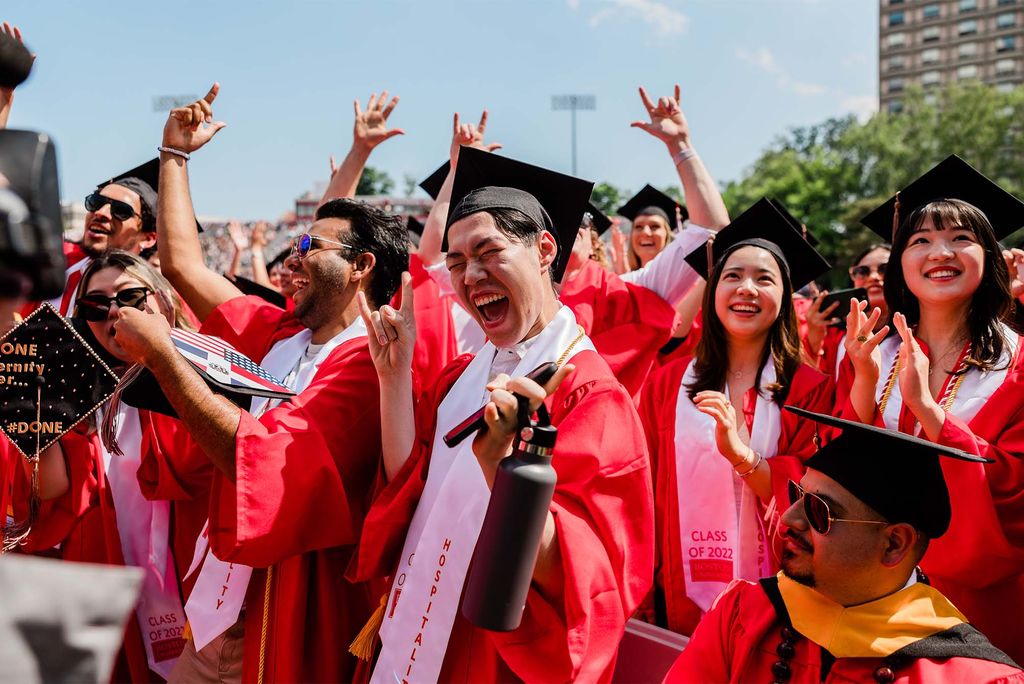 Photo of a large group of recent graduates cheering in excitement. The group of diverse students wear red gowns, black caps, and white sashes that read "Hospitality Class of 2022.". Decorations on two caps can be seen, one that shows both the American flag and the flag of the Dominican Republic, and one that reads in gold letters "One eternity later...#done"