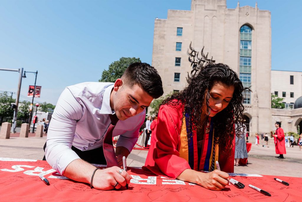Photo of two light-skinned students, a young man in a collared shirt and a woman with wavy brown hair in a red graduation robe, using sharpies to sign the class of 2022 banner.
