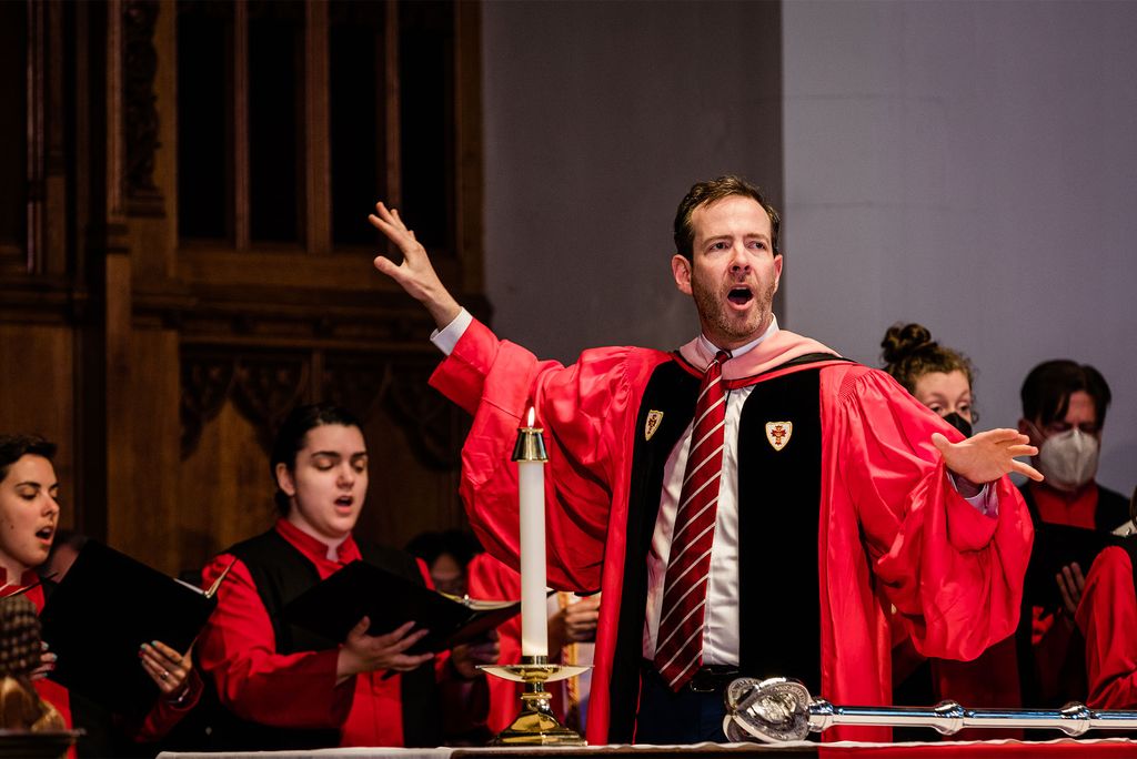 Photo of a White man with short brown hair and a short beard presumably conducting the choir at Marsh Chapel during the baccalaureate service. The conductor wears a red and black rob and robbed singers can be seen behind them.