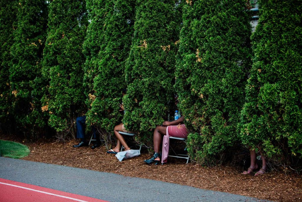 Photo of the hedges around Nickerson. Attendees brought their white chairs in between the tall, dark green hedges to get a bit of shade during commencement. In the photo, people are mostly hidden by the hedges, except for the occasional leg that appears from behind the hedges.