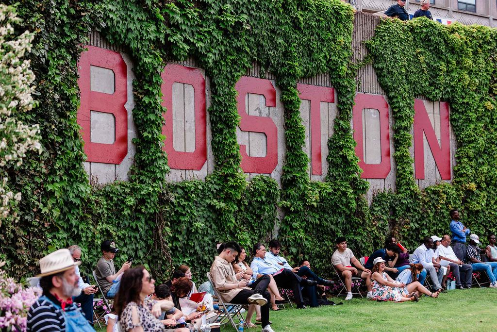 Photo taken of graduation attendees sitting in lawn chairs underneath the red Boston sign on the wall of Nickerson, which is covered by Ivy. The group wears hot weather clothing, including sun hats, shorts, and sunglasses.