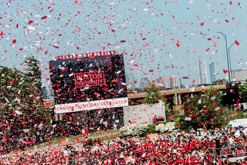 Photo: a cloud of red and white confetti rains down on a large crowd of gradation students dressed in red robes and black caps on Nickerson Field. A jumbotron seen on the field reads "Boston University, Congratulations Graduates"
