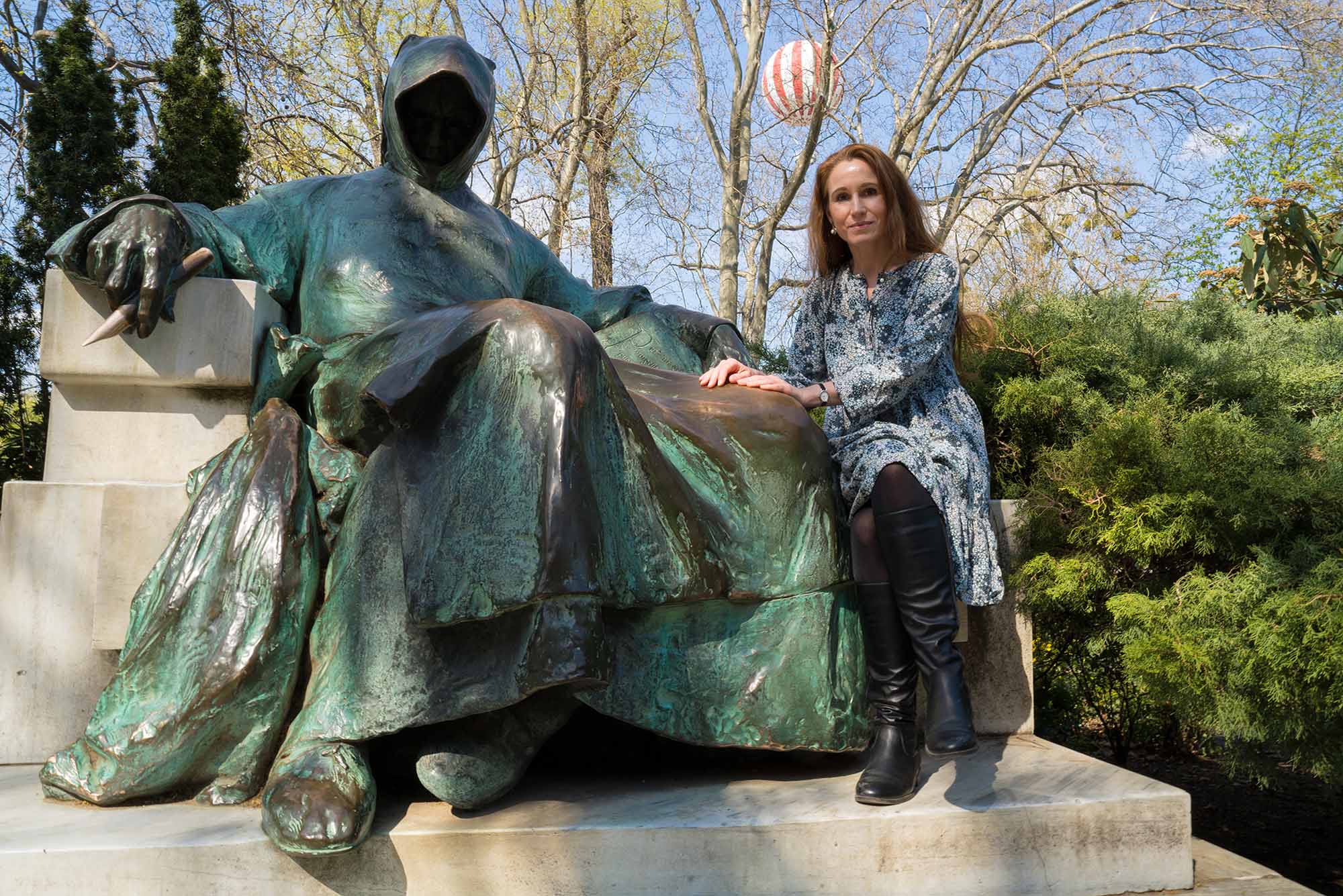 Photo of Theodora Goss in Budapest park with teal rusting bronze statue of Anonymus, a well known landmark. A white and red hot air balloon can be seen in the distance of the background, behind naked tree branches.