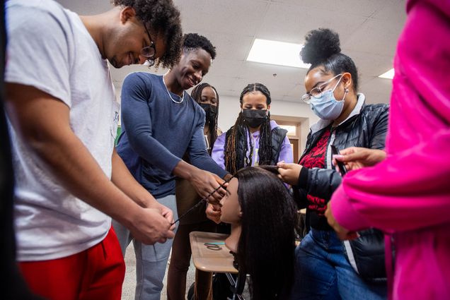 Photo of Aymen Yibrie, from left, N’Sikan Keendow, Abigail Mureithi, Axum Afework, Daniela Baez, and Naomi Boye work on their braiding during Naturally BU’s braiding demonstration at CAS. Various Black students stand around a mannequin head clipped to a desk as they practice and laugh. A few students wear masks.