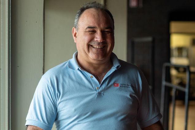 Photo of a smiling Eduino Torres standing next to a large window in the GSU. He wears a light blue collared shirt that has a small logo on the right that reads "BU Facilities Management & Operations" and jeans.