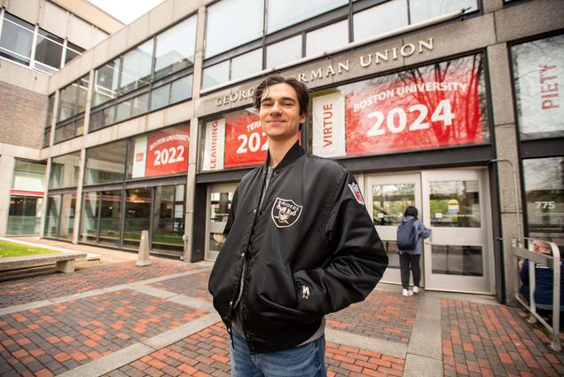 Photo of BU vlogger, Will Loggia as he stands in front of the GSU wearing an all black varsity jacket. A young, caucasian man stands with hands in pockets as he poses for a photo in front of a building where large red banners that read "Boston University 2022, Terriers 2023, and Boston University 2024".