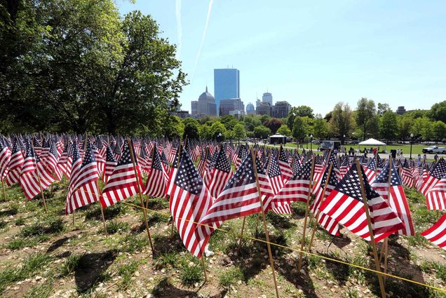 Photo of flags on Boston Common in Boston, which are placed there for Memorial Day, on a sunny day. Small American flags are planted in the ground in neat rows. in the distance tall city buildings can be seen, as well as people waling on a pathway below the flags.