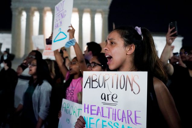 Photo of a crowd of people gathered outside the Supreme Court in Washington on the night of May 3. They protest overturning the 1973 case Roe v. Wade. Photo focuses on a young woman holding a sign mid-shout and holding a sign that reads "Abortions are Healthcare, Necessary, Justice. Keep them Legal!"