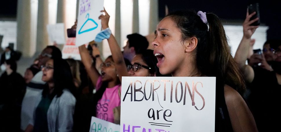 Photo of a crowd of people gathered outside the Supreme Court in Washington on the night of May 3. They protest overturning the 1973 case Roe v. Wade. Photo focuses on a young woman holding a sign mid-shout and holding a sign that reads "Abortions are Healthcare, Necessary, Justice. Keep them Legal!"