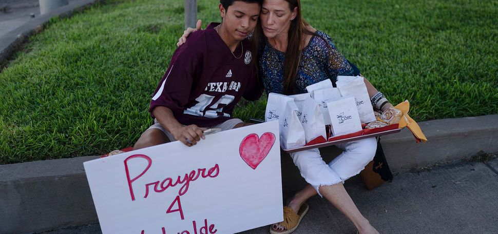 Photo of Diego Esquivel, left, and Linda Klaasson sit on curb at the edge of a green space and comfort each other as they gather to honor the victims killed in Tuesday's shooting at Robb. Esquivel is a young, Latinx tween boy who wears a crimson Texas jersey. He holds a sign that says "Prayers 4 Uvalde." Klaasson is a middle aged woman with light-brown skin and long brown hair. She closes her eyes as she gives Esquivel a side a hug. Balanced on her lap is a tray of white paper bags with presumably food in them; blue label on the bag reads "the Core" and has a logo of an apple core with a couple bites out of it.