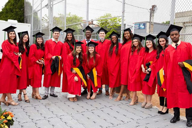 Photo of Graduate Medical Sciences master’s degree candidates before their convocation ceremony. A diverse group of young adults stand in a group wearing red caps and gowns and black caps on a sunny day.