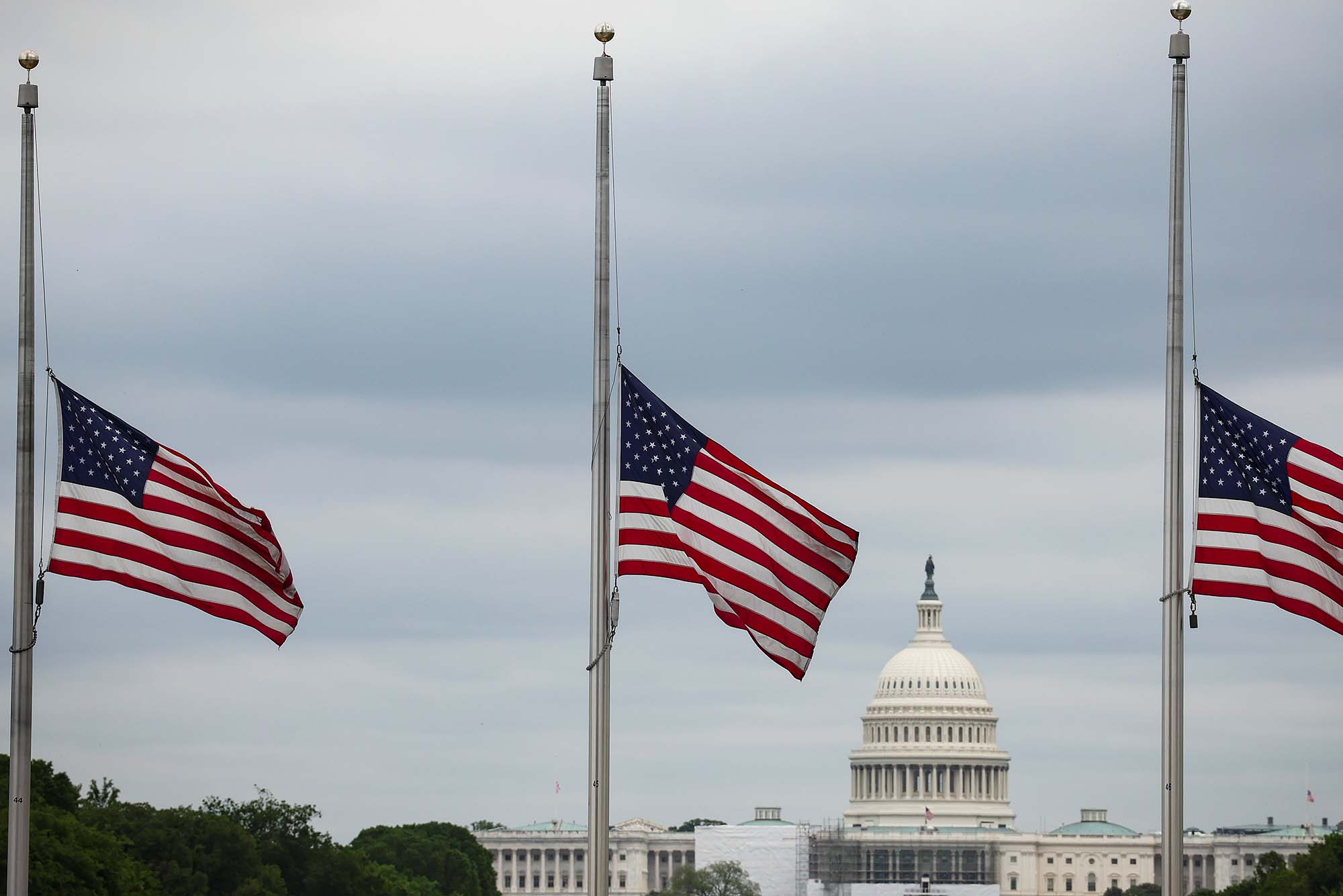 Photo of three American flags at the base of the Washington Monument flying at half staff. President Biden ordered flags to fly at half-mast through next Monday as the nation passes the 1 million mark of COVID deaths. The sky in the background is cloudy and gray, and the top of the white, capital building is seen in the background at bottom.
