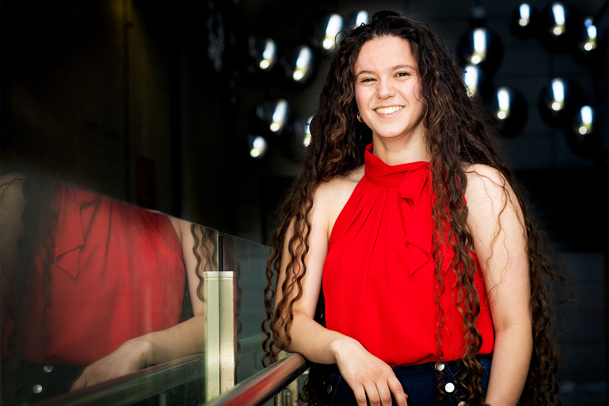 Amanda Berke standing smiling in a red top next to a mirror in front of a dark background