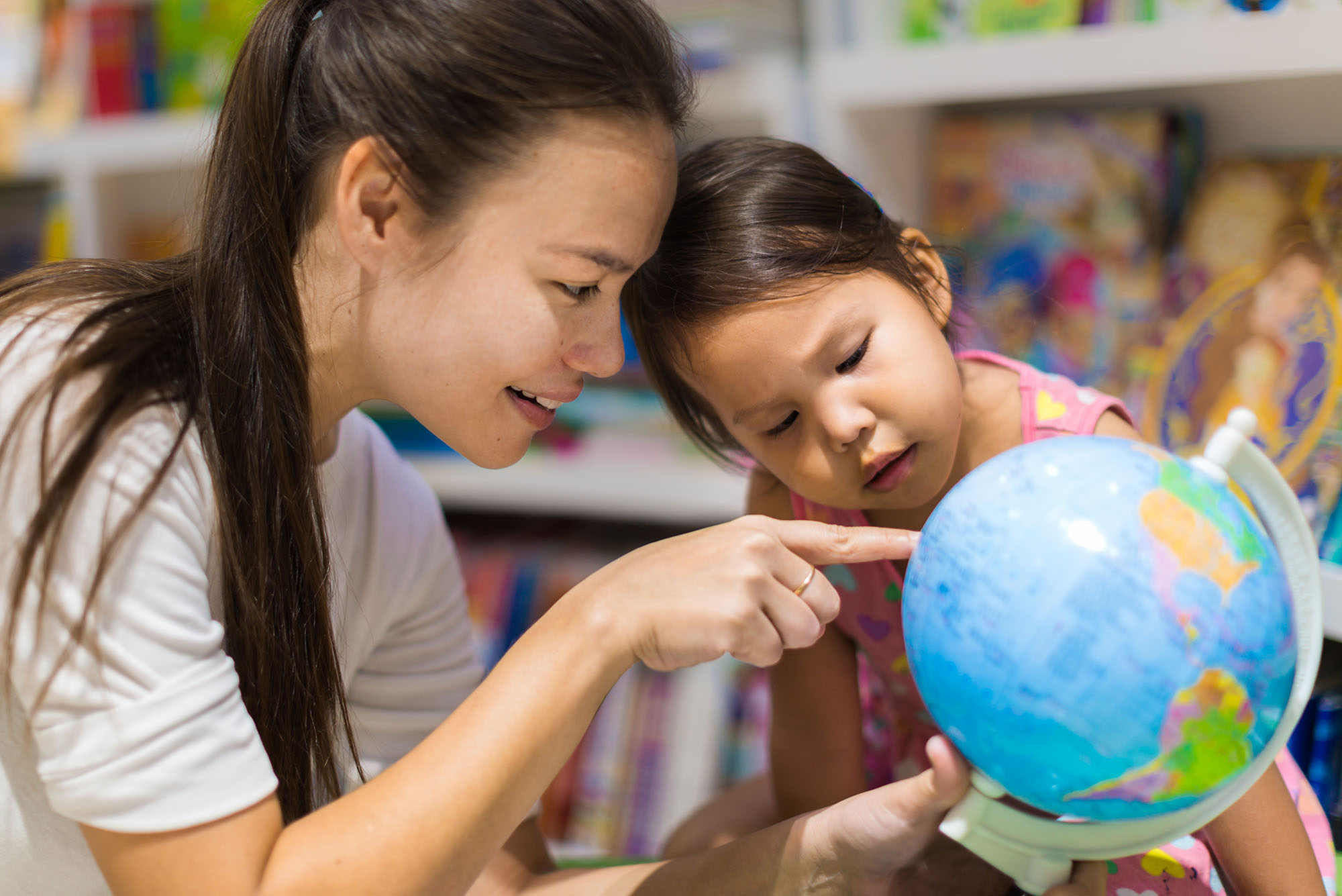 Photo of an Asian parent pointing something out to her child on a small globe in a library bookstore.