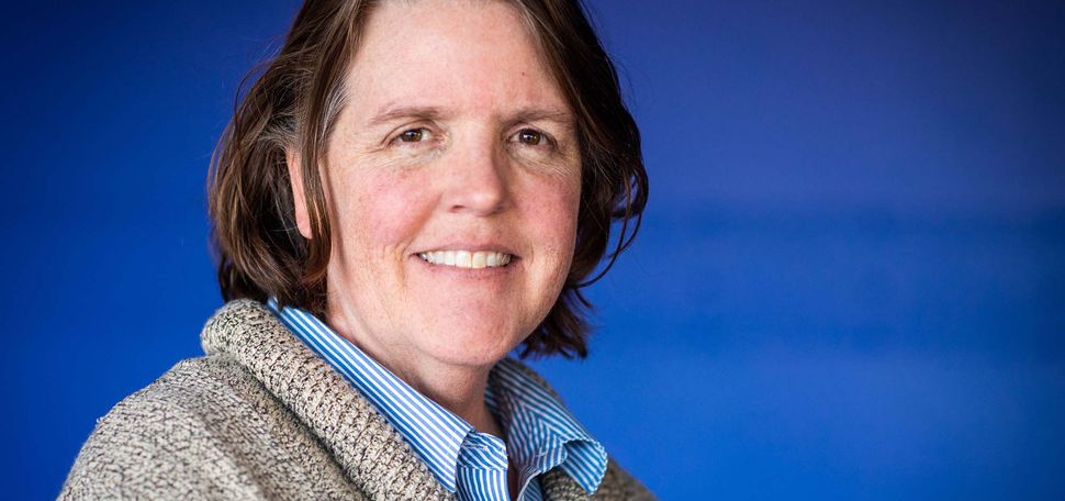 Photo of Metcalf winner Leslie Dietiker, an older white woman with short brown hair with some gray streaks in it. She smiles, and wears a thick, gray sweater and striped blue shirt. She stands in front of a blue background.