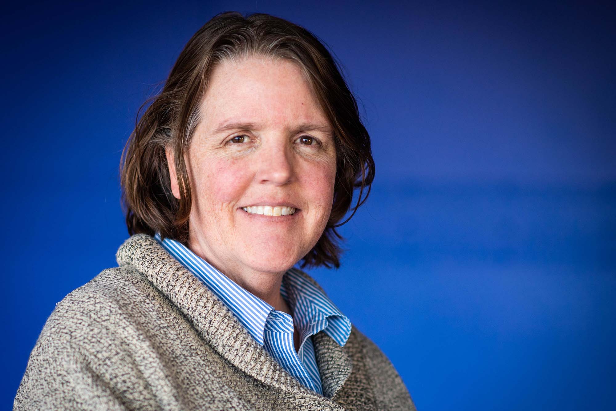 Photo of Metcalf winner Leslie Dietiker, an older white woman with short brown hair with some gray streaks in it. She smiles, and wears a thick, gray sweater and striped blue shirt. She stands in front of a blue background.