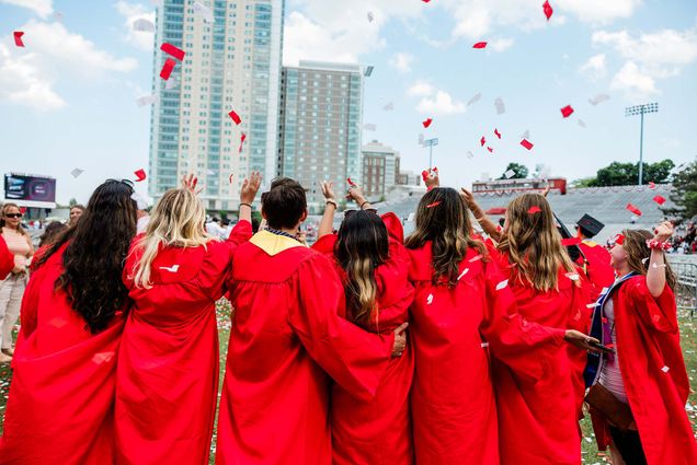 photo taken behind six graduates at the end of the commencement ceremony. they wear their red gowns and throw confetti in the air. warren towers is seen in the background, as well as a light blue sky with a few small clouds. Most of the students have long blonde or brown hair.