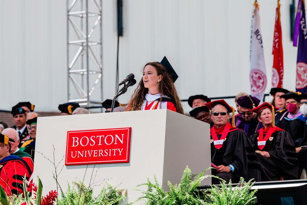 0457: Photo of Amanda Berke (CAS’22, GRS’22), a White woman with very long brown wavy hair, who wears a black mortar board and red gown. She is seen mid-speaking, smiling, at a large white podium with a red "Boston University" sign. A floral arrangement is seen in the foreground; behind her, honorary degree recipients, faculty members.