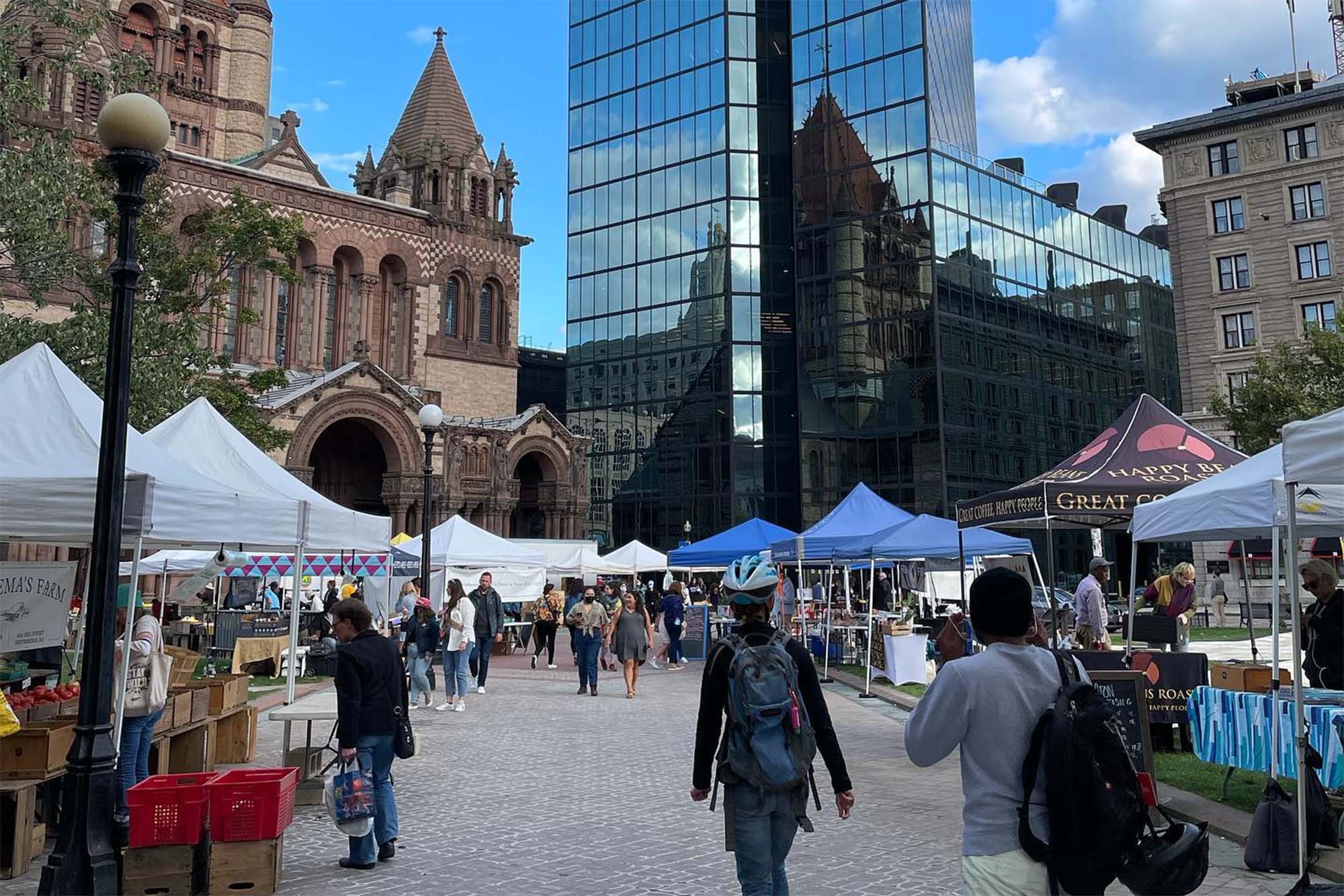 Photo of the Copley Square Farmer's market on a sunny day. Shoppers are seen between the lines of pop-up tents at left and right. Trinity Church, with is brown stone facade, is seen at left. The sky is blue with one puffy cloud.