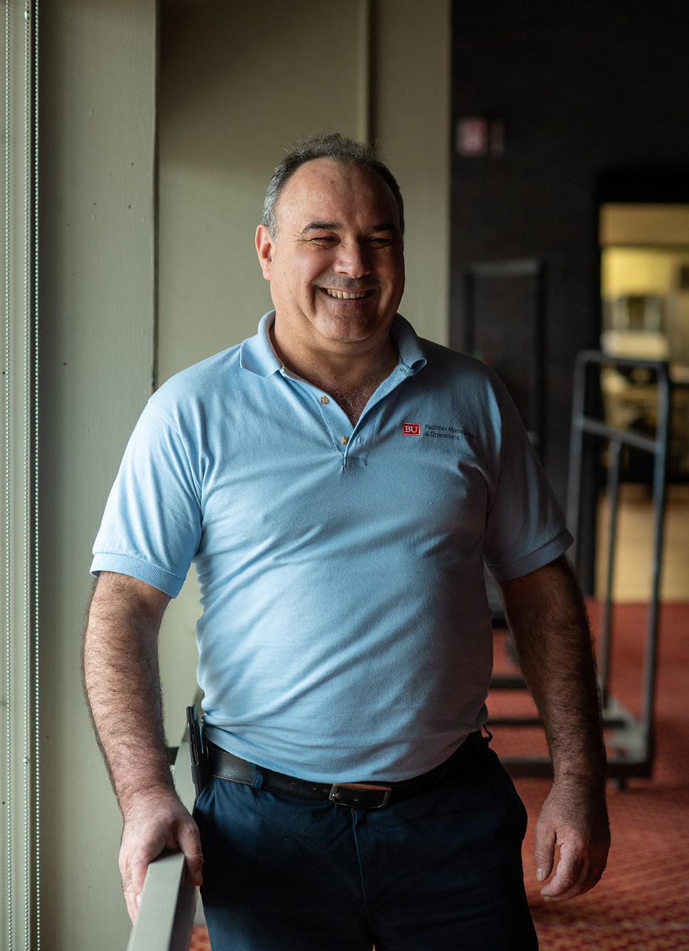 Photo of a smiling Eduino Torres standing next to a large window in the GSU. He wears a light blue collared shirt that has a small logo on the right that reads "BU Facilities Management & Operations" and jeans.