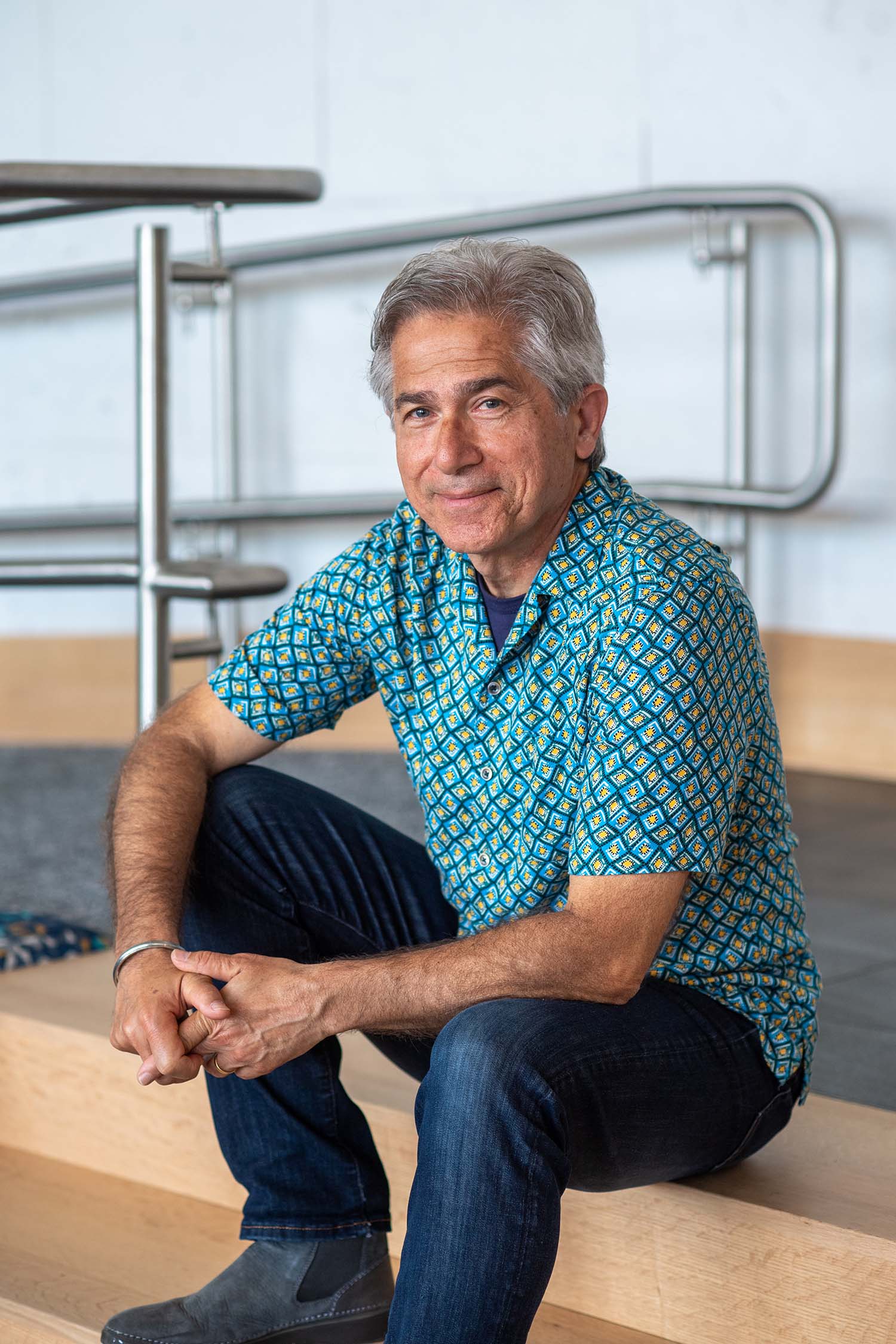 Photo of Armenian-Iranian Albrik Avanessian (MET’22). He is in his 60s, has short gray hair, and has a sincere smile. He wears a blue and white patterned shirt and dark jeans, and sits on a wooden step, resting his arms on his knees.