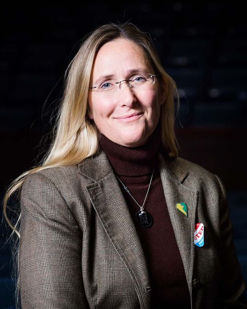 Photo of Scarlet Lewis, a middle-aged White woman with long blonde hair, and small glasses. She smiles and wears a tweed blazer and maroon turtle neck. A "I voted" sticker is on her blazer. She poses for this portrait in front of a black background.