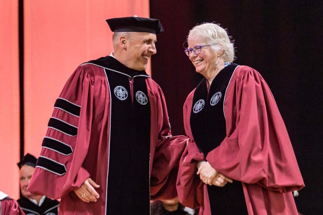 Photo of Colgate University President Brian W. Casey with BU provost Jean Morrison. Both wear pale red robes, Casey's has black stripes down the arms, and they both wear a black sash with a white logo embroidered. Casey is an older White man who smiles and wears a black hat as he shake Jean Morrison's hand. Morrison is an older White woman with glasses who smiles. Both appear on stage.