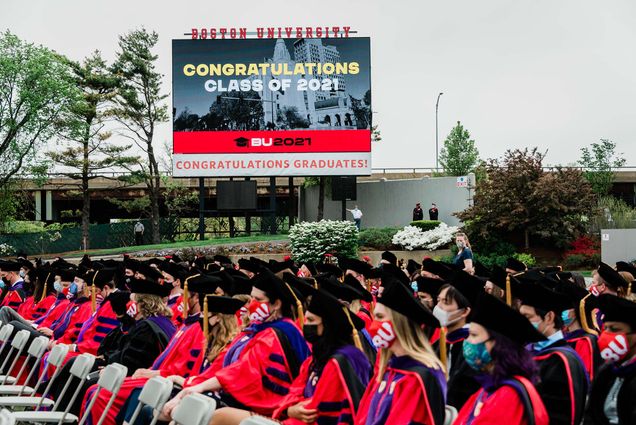 Photo of students sitting on Nickerson field during the 2021 commencement. They wear black graduation caps and red gowns. Behind them, the large jumbotron screen is seen. It reads "Congratulations class of 2021" on screens, and below, a white banner reads "Congratulations graduates!"