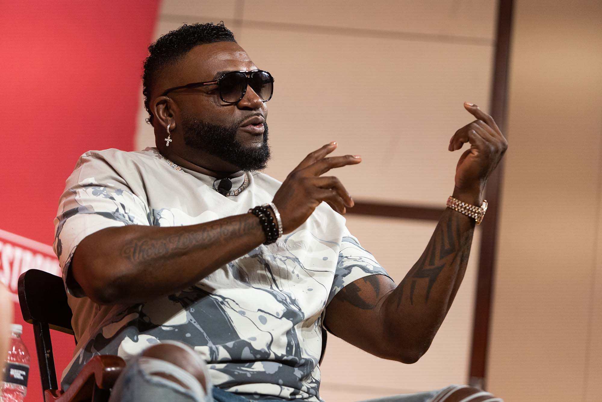 Ortiz on stage during his Q&A at Questrom. Ortiz, a middle-aged Black man, is seen seated from the side, and gestures with his hands; he wears ripped jeans, a gray graphic tee and sunglasses.