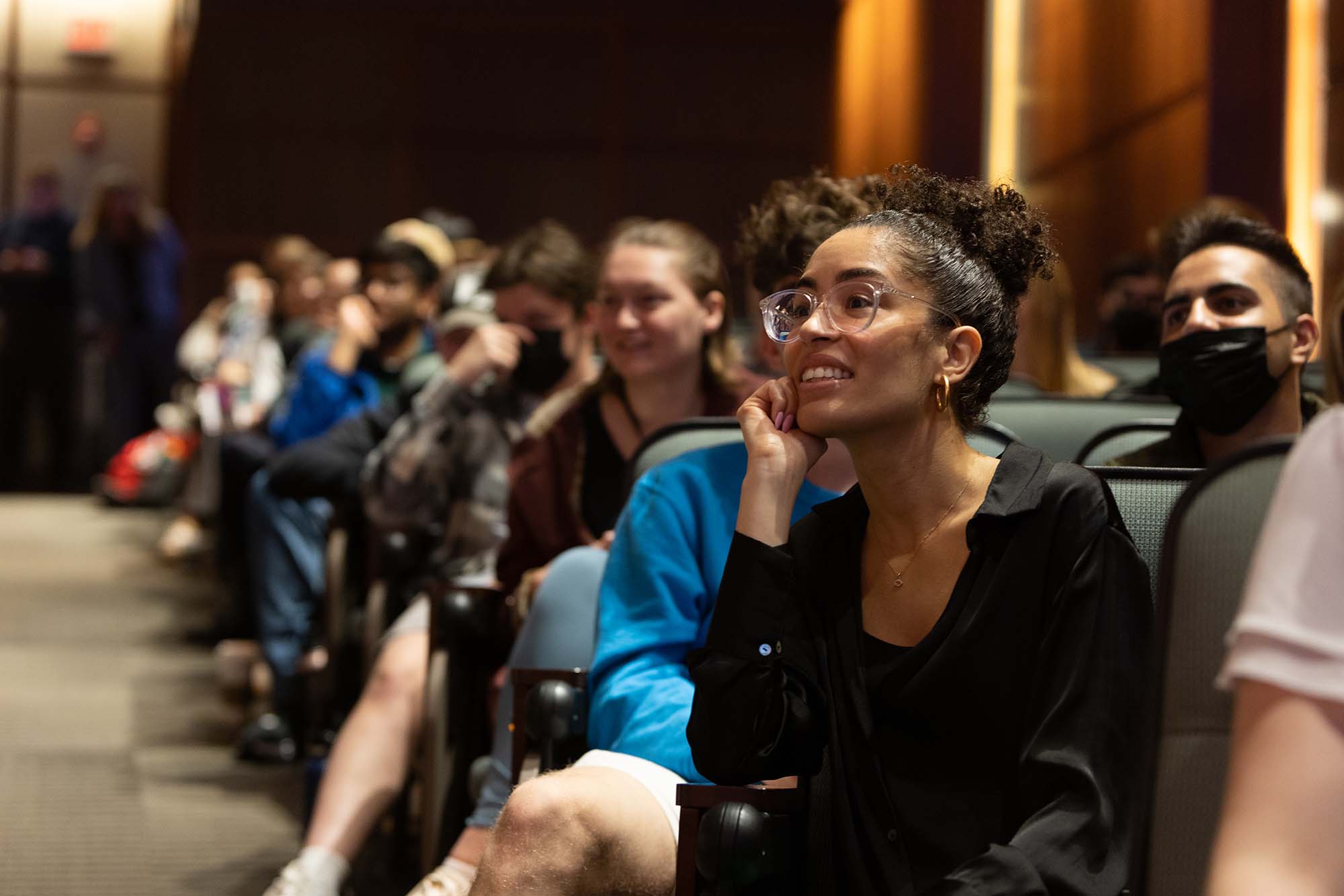 An audience member in the Questrom auditorium looks toward the stage during Ortiz's Q&A. The young woman Black has glasses, wears Black, and rests her hand on her chin as she listens. The auditorium rows behind her a filled.