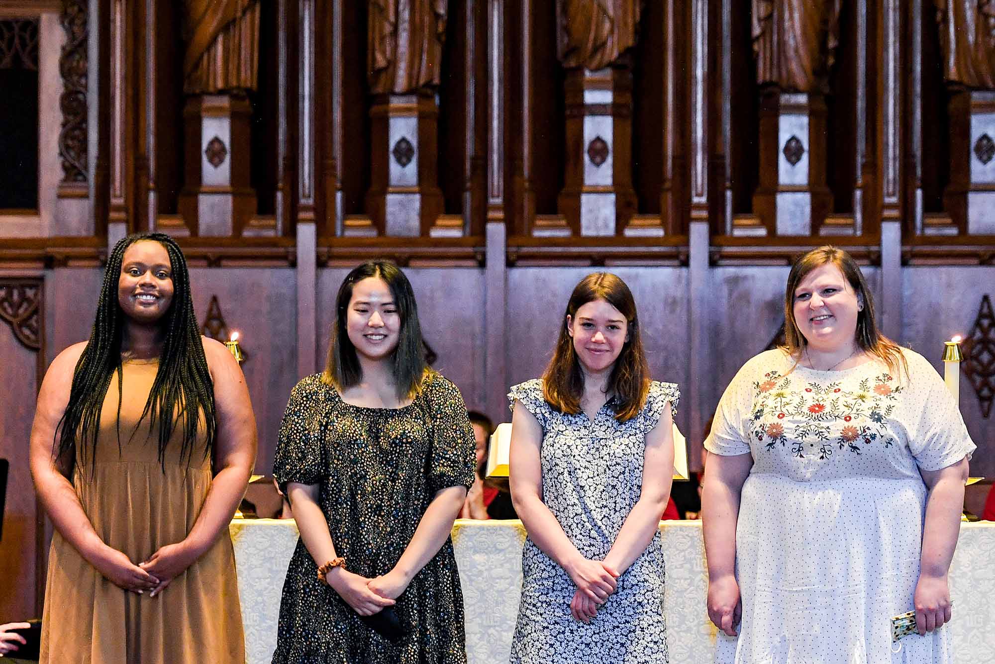 Photo of four This I Believe student speakers standing in front of the altar at Marsh Chapel: Kendall Lowe (CAS'22), Yeeun Jang (CAS'22, Anna Jensen (CAS'22), and Chloe McLaughlin (STH'22). Lowe is a young, Black woman with long braids, who smiles, clasps her hands and wears a tan dress. Jang is a young AAPI woman who wears a black floral dress, has shoulder-length black hair and looks down and smiles. Jensen is a White woman wearing a blue and white floral dress and clasps her hands as she smiles, she has dirty blonde hair. McLaughlin is a White. woman wearing a white dress with polkadots and a floral pattern. She smiles, looks to her right and has shoulder length dirty blonde hair. The brown, wood-carved wall is seen behind them.