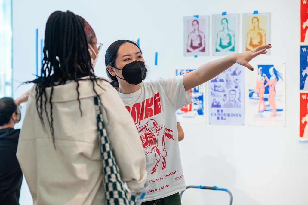 Photo of Mary Yang, CFA assistant professor of graphic design, gesturing with her left hand, as she helps her students hang their work in preparation for this year’s BFA Thesis show. A student of color is seen in the foregroud looking towards Yang. Behind Yang, a series of colorful prints, portraits of a man, hang.