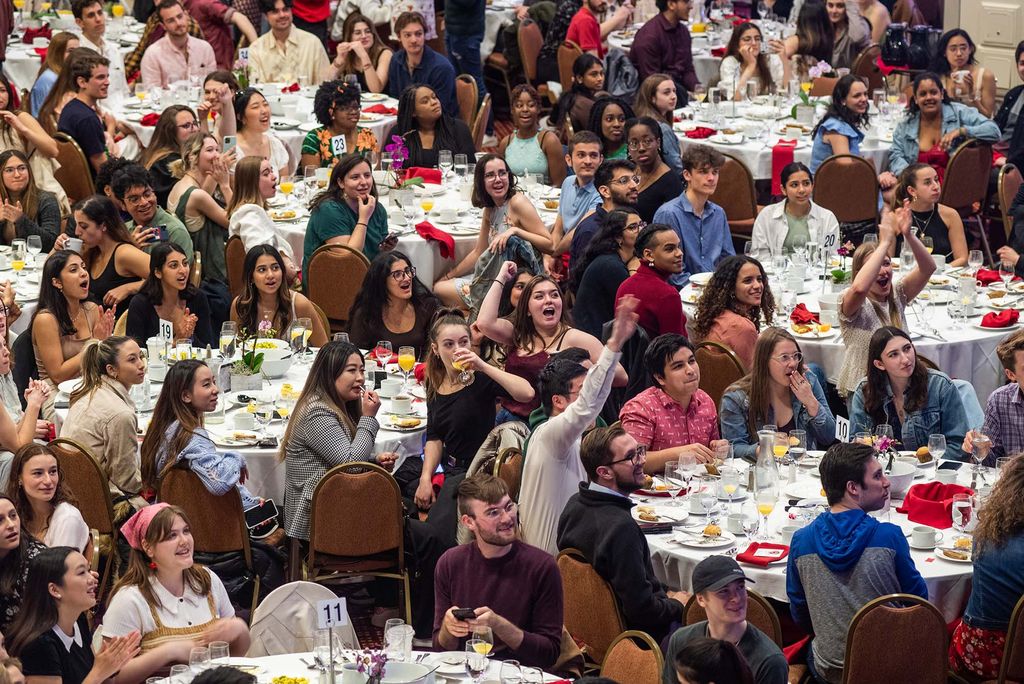 A diverse group of students, all seated at large round tables with white table clothes, some eating and drinking, others reacting and cheering as they watch a slideshow looking back on their college experience during senior class breakfast at the GSU’s Metcalf Ballroom May 5. 