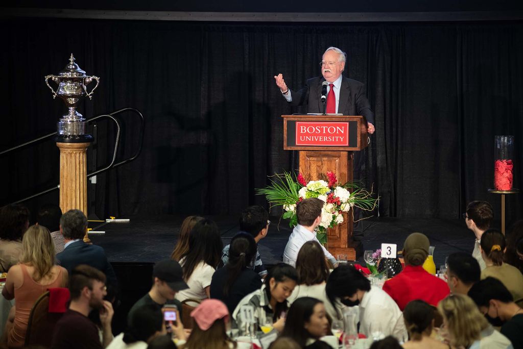 Photo of President Robert A. Brown addressing the class of 2022 during senior class breakfast at the GSU’s Metcalf Ballroom May 5.  He is an older White man who wears a suit and red tie, and gestures with his right hand as he speaks at a wooden podium with a red Boston University sign on it. At left, a silver trophy is seen, at right, a clear container of red tickets. Seniors are seen from behind as they listen to Brown speak.