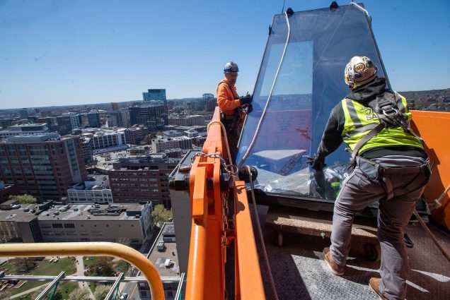 Photo of two construction workers at the edge of one of the floors of the Center for Computing and Data Science. The men wear hard hats and help lower what looks to be a piece of glass that has been raised by a crane. Behind them, the COM lawn and Boston city skyline is seen.