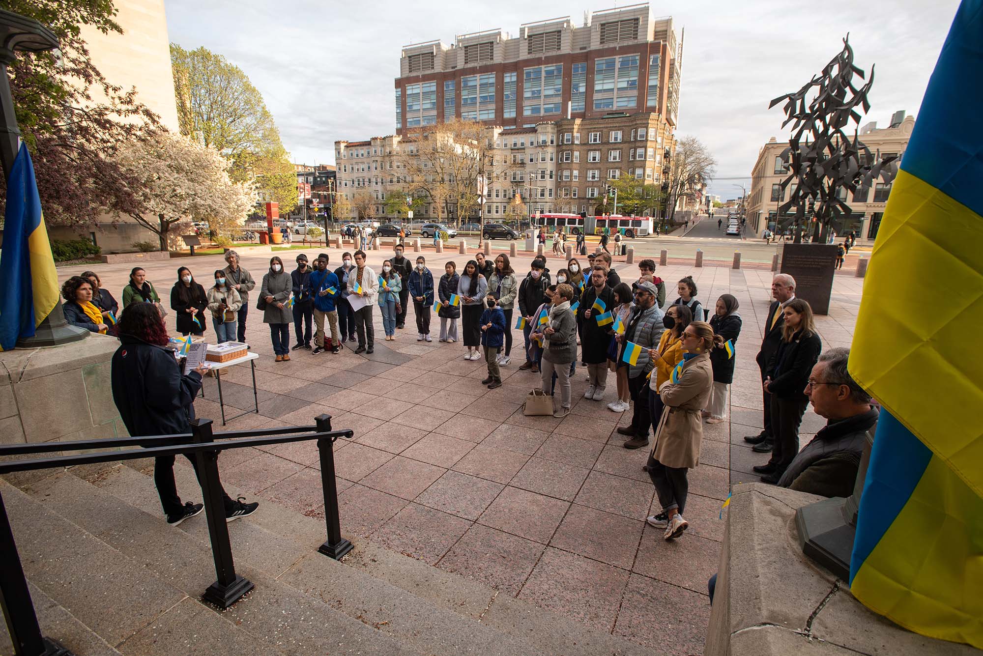 Wide shot of a group of people gathered on Marsh Plaza. At left and right of the steps, large blue and yellow Ukraine flags fly. Some members of the group are masked, many hold small Ukrainian flags. It's an overcast day.