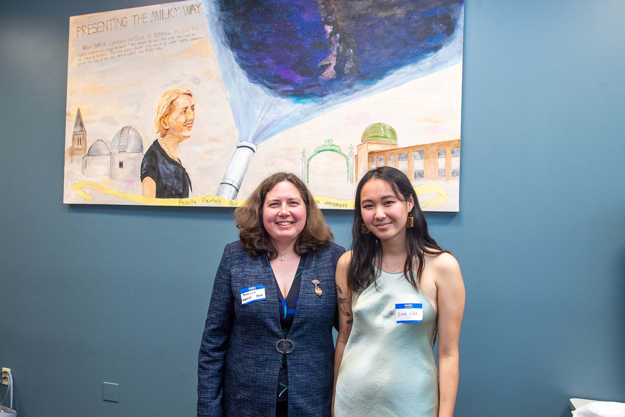 Photo of Boston city councillor Kenzie Bok, left, with mural artist Zora Che (CAS’22), standing in front of Che's mural honoring BU alum Priscilla Fairfield Bok on May 13 in CAS. Bok, a middle aged white woman with brown hair, wears a blue formal jacket and smiles. Che is a young AAPI woman with long black hair who wears a pale green dress, square dangling earrings, and smiles. The mural is on a blue teal wall, and features and image of Priscilla Fairfield Bok with a telescope that appears to project an image of the milky way.