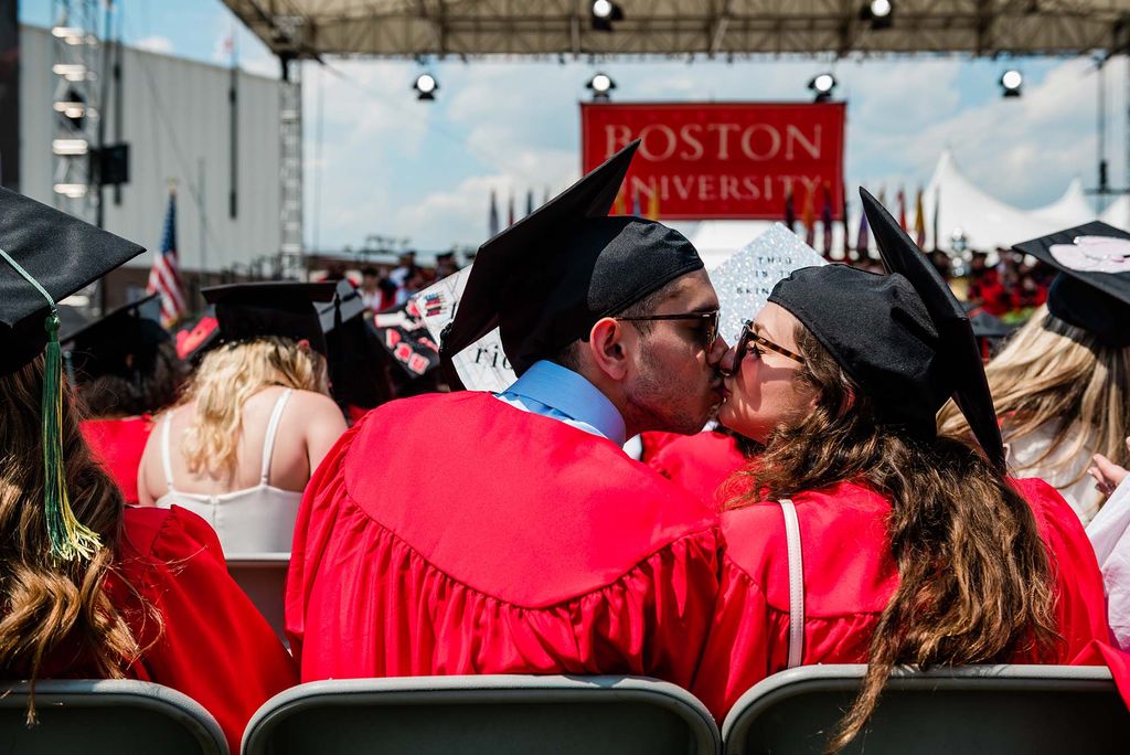 0443: Photo of a young couple, a young man and young woman, kissing during the commencement ceremony. They both wear their black mortarboards, red gowns, and sunglasses. The photo is taken from behind them, and in the background you can see the commencement stage and a large red sign that reads "Boston University."