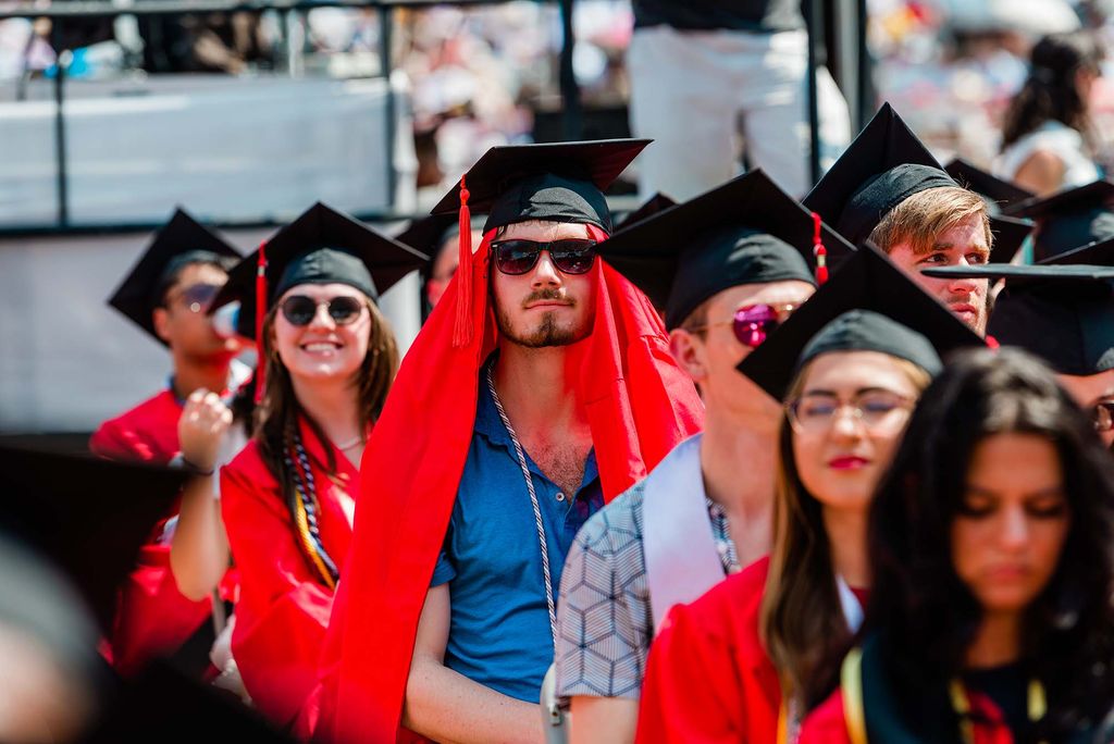 Photo of a student at commencement wearing his rob on his head underneath his mortarboard. He wears sunglasses and a blue button down and appears to be wearing his gown on his head to beat the heat. Other students are seen sitting on Nickerson smiling during the ceremony.