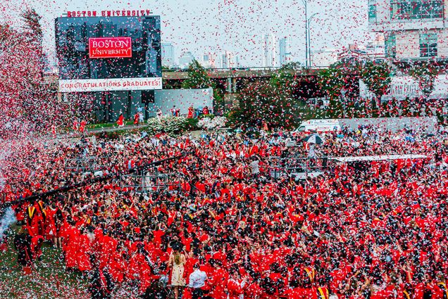 Photo taken of red and white confetti canons going off at the end of commencement. Graduates can be seen on on the field in their red gowns and black mortarboards, and the jumbotron is seen at left.