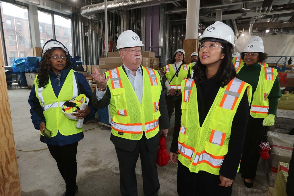 Photo Michelle Wu, right, and Bob Brown, left, as well as other folks, wearing bright yellow vests and white hard hats. Bob Brown is gesturing with his right hand as Michelle Wu looks up at presumably construction happening at the Data Sciences Center. Boxes and construction materials are seen behind them.