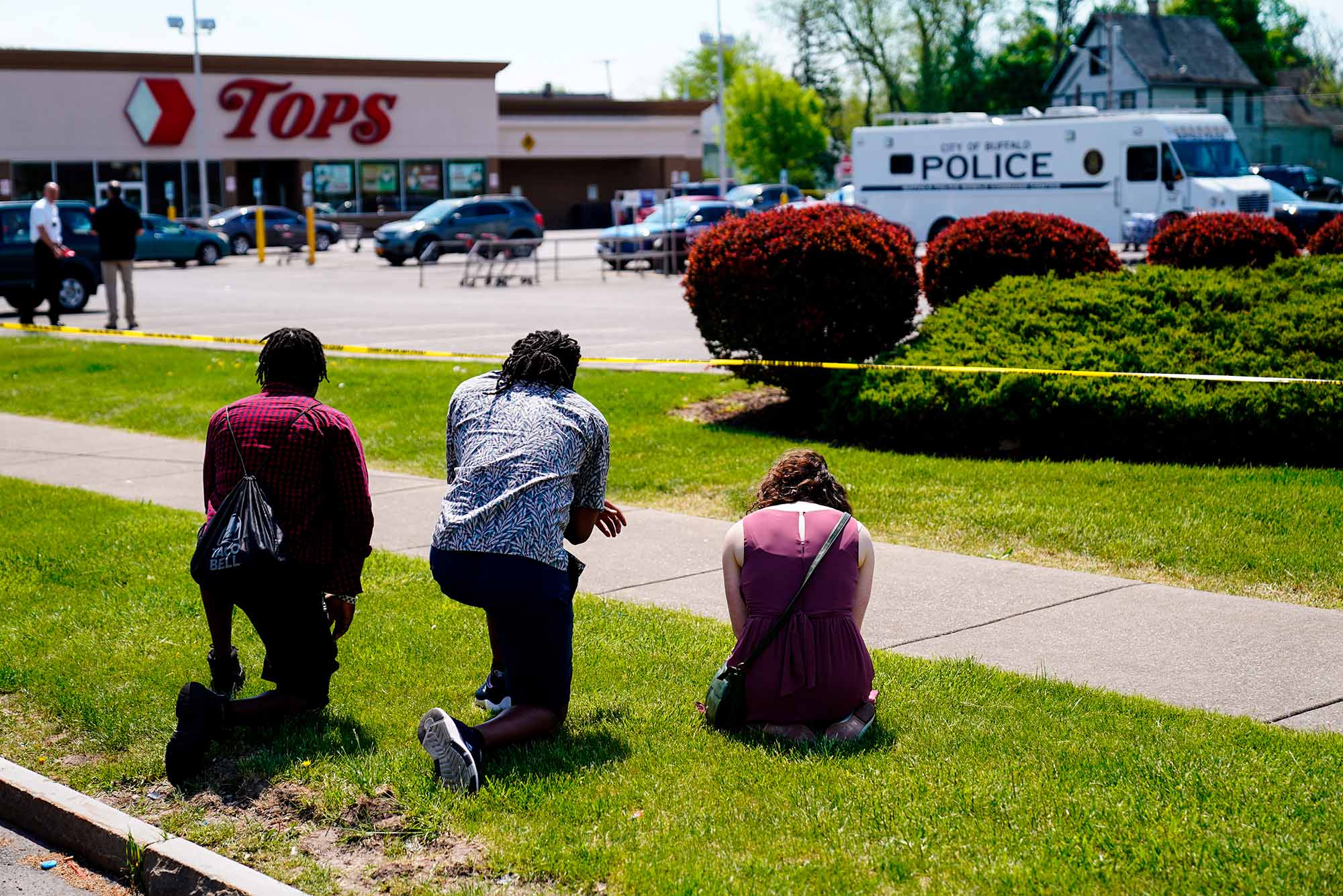 Photo of people praying, two young Black men and one White women, outside the scene of Saturday's shooting at a supermarket, in Buffalo, N.Y., Sunday, May 15, 2022. Behind them, the TOPS supermarket sign is seen across the parking lot. Also in the parking lot a parked police van.