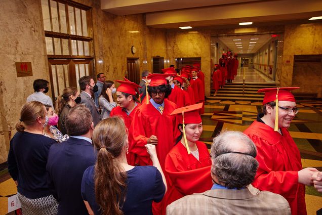 Photo of a line of young, Boston University Academy students fist-pumping their teachers and school staff as they process into their graduation. They a diverse group that wears red caps and gowns with gold tassels. The floor has a geometric tile pattern and the hallway is lit with yellow-ish light.