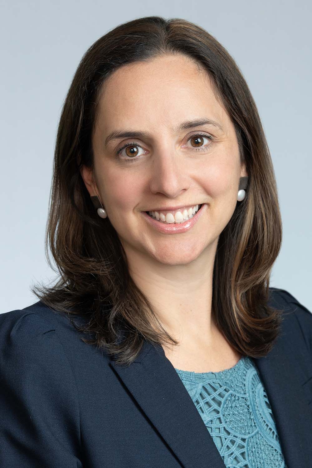 Portrait of Jen Green, a White woman with shoulder length brown hair who wears a blue blazer and light blue blouse. She smiles and also wears pearl earrings.